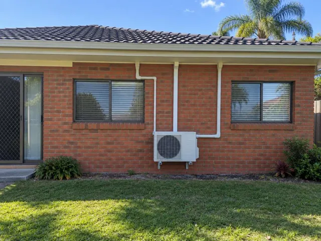 Wall-mounted outdoor air conditioning unit with white pipe covers on a brick house exterior in Carrara